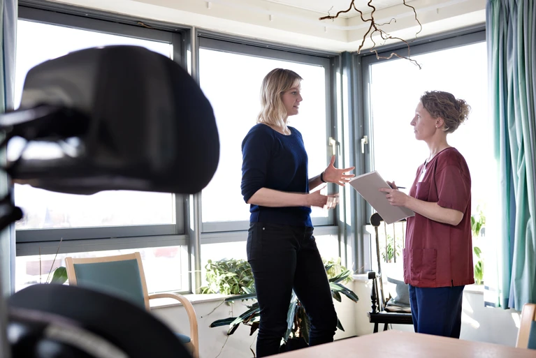 Totalskud af to kvinder, Ditte og Helle, som står og taler med hinanden foran vinduerne i et lokale. I forgrunden ses dele af en kørestol med nakkestøtte.