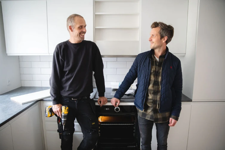 To mænd står i et køkken. Den ene har en i arbejdstøj og med en skruetrækker i hånden. De ser venligt på hinanden.