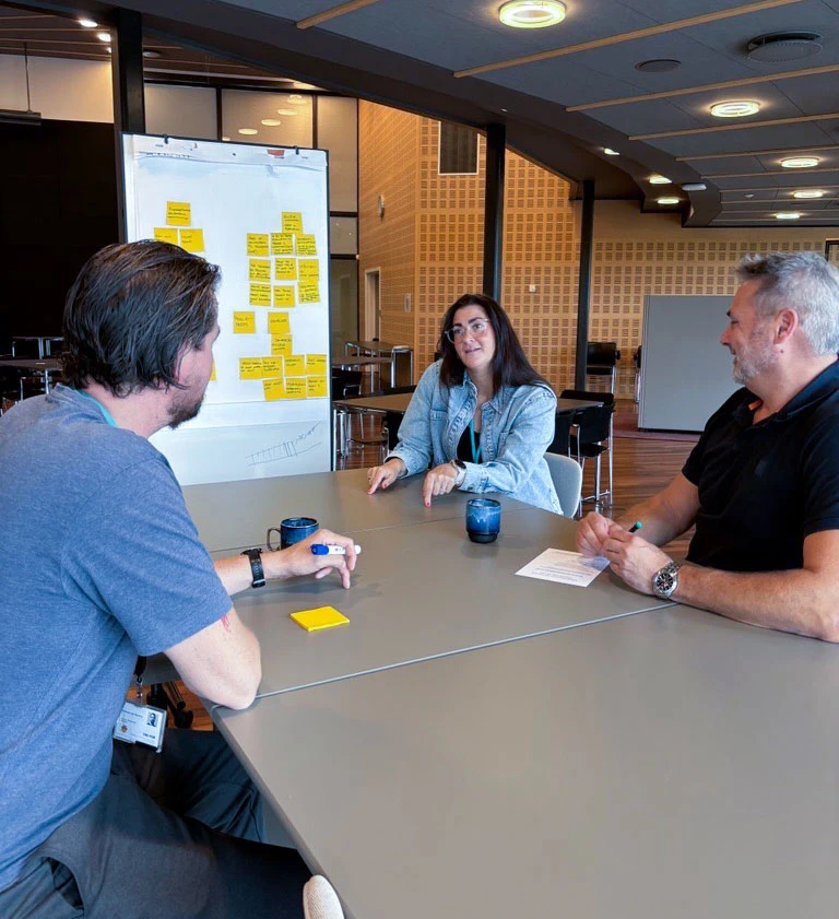 Tre mennesker sidder i et mødelokale om et bord og taler med hinanden. Der er en tavle med gule post-its på bag dem.