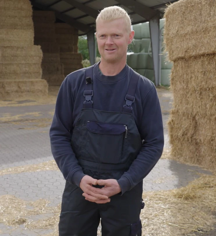 Landmand står på gårdsplads med halmballer i baggrunden.  