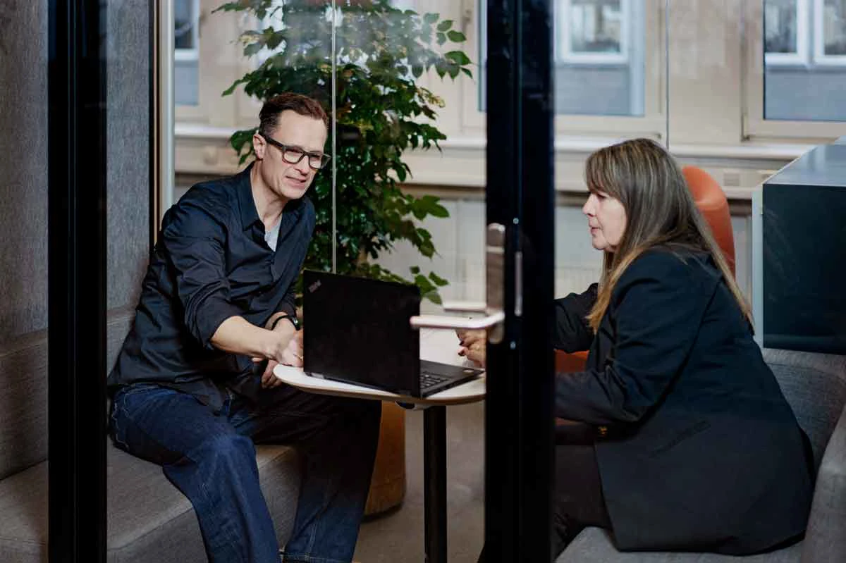En kvinde og en mand sidder ved en computer på et kontor og snakker.